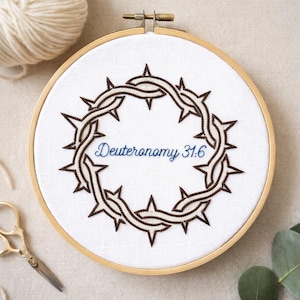 Könnte beinhalten: Ein Stickrahmen mit einem braunen Dornenkranz-Design auf weißem Stoff. Der Text "Deuteronomium 31:6" ist blau gestickt. Ein Holzrahmen hält den Stoff, mit einem Wollknäuel und einer Schere im Hintergrund.