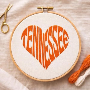May include: An embroidery hoop with a heart-shaped design that says "Tennessee" in orange thread. The hoop is made of wood and the fabric is white. Gold scissors and orange and white thread are also in the image.