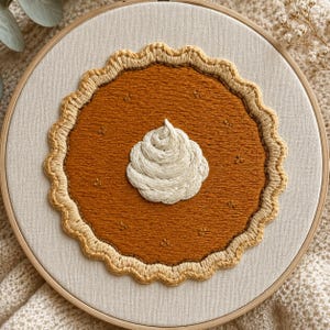 Puede incluir: Un bastidor de bordado circular con un diseño cosido a mano de una tarta de calabaza sobre tela color hueso. La tarta presenta un relleno naranja oscuro, una corteza festoneada color canela claro y una nata montada blanca central.