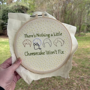 Może przedstawiać: Haft na tamborku z napisem „There's Nothing a Little Cheesecake Won't Fix”. Projekt przedstawia haftowane obrazy trzech fryzur damskich i okularów. Tło to jasnobeżowa tkanina z żółtymi haftowanymi kropkami.