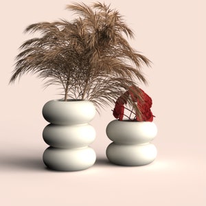 Puede incluir: Dos macetas blancas con forma de esferas apiladas, una grande y otra pequeña, cada una con plantas secas. La maceta más grande contiene una planta marrón alta y plumosa. La maceta más pequeña contiene una planta roja y frondosa.