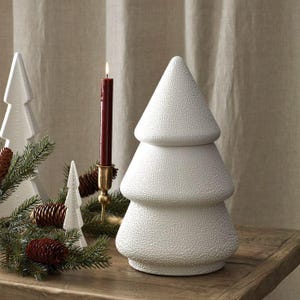 Puede incluir: Un árbol de Navidad de cerámica blanca con superficie texturizada, exhibido sobre una mesa de madera. Una vela burdeos encendida en un portavelas de latón y otros árboles decorativos y piñas crean una escena festiva navideña.