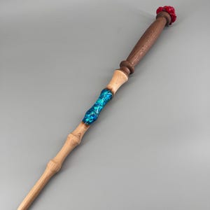 May include: A handcrafted wooden wand with a light brown handle and a dark brown grip. The wand features a vibrant blue resin section and a red rose-shaped tip. The wand is a unique fantasy item.