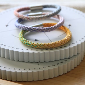 May include: Three braided bracelets in different colours, including pink, purple, and green, with gold accents, are stacked on top of each other on a white circular tool with numbers.