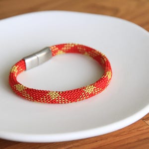 May include: A red and gold braided bracelet with a silver clasp. The bracelet is on a white plate.
