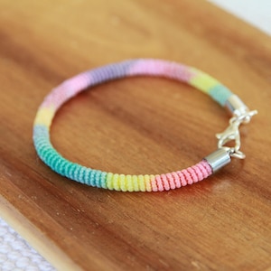May include: A colorful bracelet with a rainbow gradient of pink, yellow, green, and purple. The bracelet is made of a woven material and has a silver clasp. The bracelet is displayed on a wooden surface.