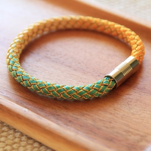 May include: A braided bracelet with a gold and green color gradient. The bracelet has a silver clasp.