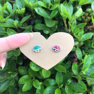 May include: A pair of earrings featuring a green and red turtle shell design. The earrings are displayed on a heart-shaped piece of cardboard.