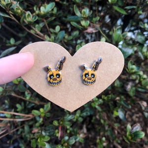 May include: A pair of yellow and brown stud earrings featuring a cartoon character with a wide smile and large ears. The earrings are displayed on a heart-shaped piece of cardboard.