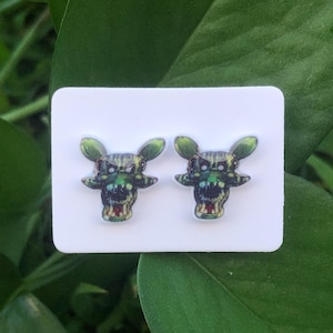 May include: A pair of stud earrings featuring a green and black monster face design. The earrings are on a white card, set against a backdrop of green leaves. The monster face has sharp teeth and pointed ears.