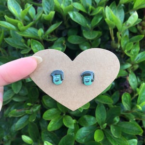 May include: A pair of stud earrings featuring a cartoon character's face with a green complexion, black hair, and a blue shirt. The earrings are displayed on a heart-shaped piece of cardboard against a backdrop of green foliage.