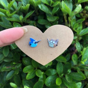 May include: A pair of cartoon character stud earrings. One earring features a blue bird with a black beak, and the other depicts a brown and tan character. The earrings are displayed on a heart-shaped cardboard backing.