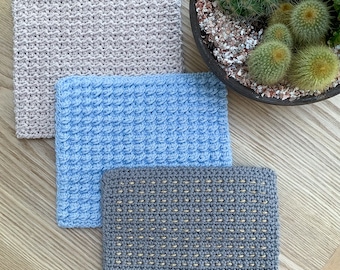 May include: Three crocheted squares in different colors and patterns. The top square is light pink, the middle square is light blue, and the bottom square is gray with gold thread.