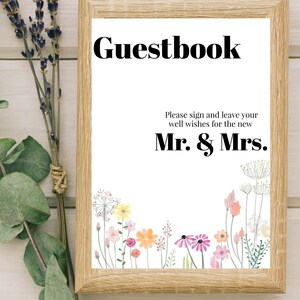Puede incluir: Un marco de madera con fondo blanco y el texto "Guestbook" en negro. El texto "Please sign and leave your well wishes for the new Mr. & Mrs." también está en negro. La parte inferior del marco está decorada con una fila de flores de colores.