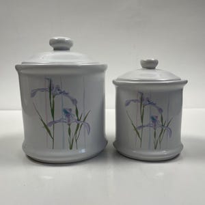 Op de afbeelding: Twee witte keramische potten met deksels. Elke pot heeft een bloemenontwerp van paarse irissen met groene bladeren en verticale strepen in roze en blauw. De potten hebben verschillende maten.