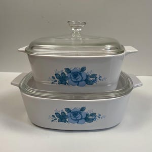 Puede incluir: Dos fuentes blancas para horno con tapas de cristal. Cada fuente presenta un diseño floral azul. La fuente superior es más pequeña y se asienta sobre la fuente más grande. Las tapas tienen pomos de cristal transparente.