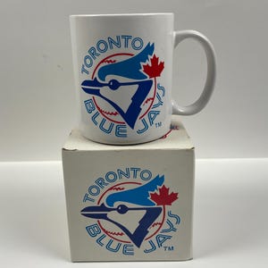 Può includere: Tazza in ceramica bianca con il logo dei Toronto Blue Jays. Il logo include una testa di ghiandaia azzurra, una foglia d'acero rossa e le parole "TORONTO BLUE JAYS" in un design circolare. La tazza è su una scatola abbinata con lo stesso logo.