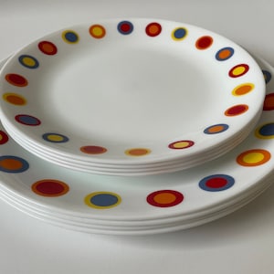Puede incluir: Un juego de platos de cena de cerámica blanca con un patrón de lunares coloridos. Los platos están apilados uno encima del otro, con el plato superior con un patrón de lunares rojo, amarillo y azul.