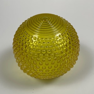 May include: A spherical yellow glass light fixture shade featuring a textured hobnail pattern across its surface and a series of concentric circular ridges at the top. The glass is translucent, allowing light to pass through the yellow material.