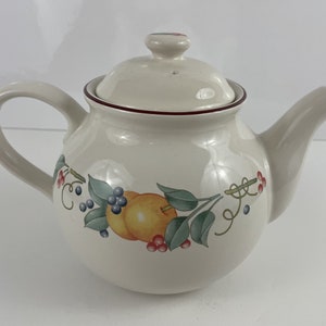 Op de afbeelding: Een witte theepot van keramiek met een rode rand en een fruitontwerp. Het ontwerp bevat sinaasappels, bladeren en bessen in tinten rood, groen, blauw en geel.