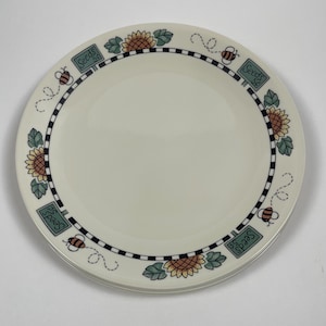 Puede incluir: Un plato de cena de cerámica blanca con un borde a cuadros blanco y negro. El borde está decorado con girasoles, abejas y hojas verdes. El texto "Seeds St." se repite alrededor del borde.