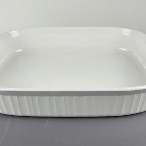 May include: A white, rectangular baking dish with a slightly curved rim and vertical ribbed detailing on the sides. The dish is empty and sits on a gray surface. This kitchenware is designed for baking and serving.
