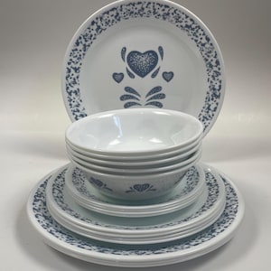Puede incluir: Un juego de vajilla Corelle blanca con un patrón moteado azul y blanco y un diseño de corazón azul. El juego incluye cuencos, platos y platillos.
