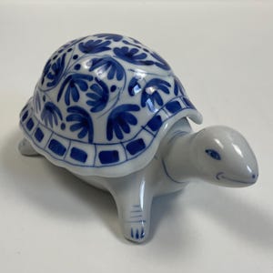 May include: A ceramic turtle figurine with a white body and a blue and white patterned shell. The shell features a floral design. The figurine is on a white surface.