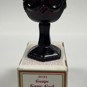 May include: A dark red wine goblet from the Avon Cape Cod Collection. The glass features a detailed, textured pattern and a black stem. The box reads "Wine Goblet" and "Fragrance Candlette Holder".
