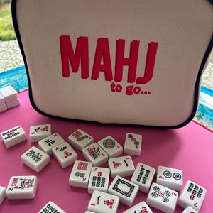May include: A beige rectangular zippered case with black piping and the red embroidered text MAHJ to go... sits behind a scattered collection of white mahjong tiles featuring various black, red, and green symbols on a bright pink surface.