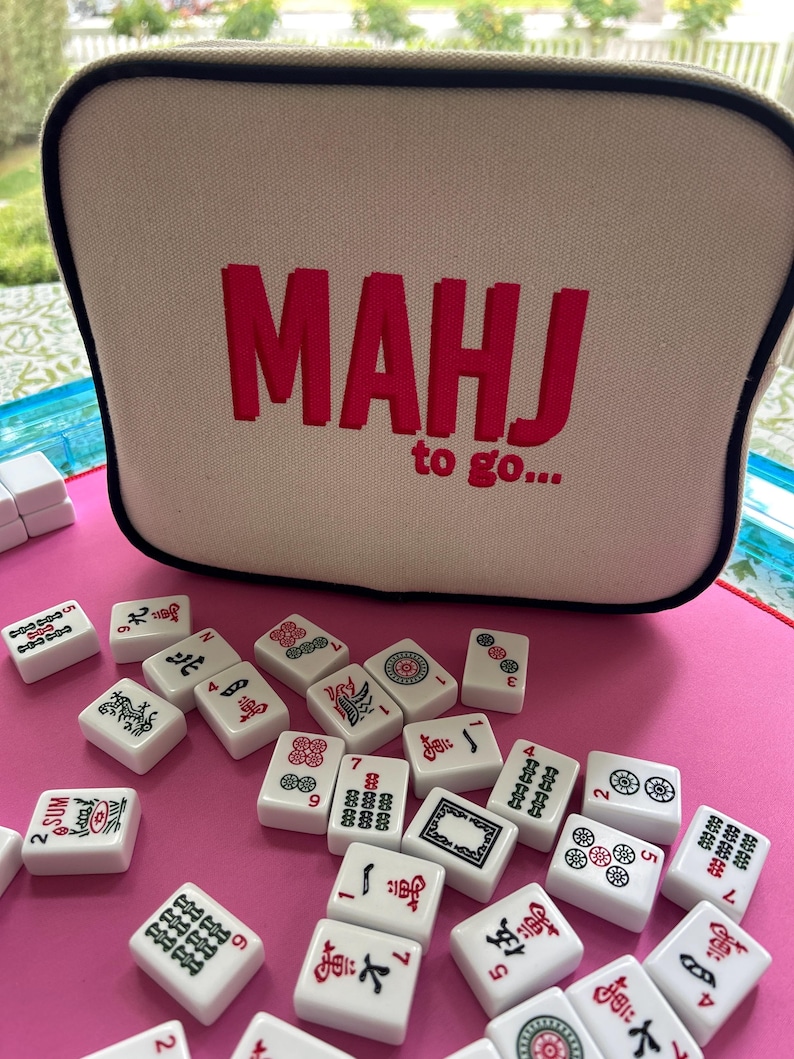 May include: A beige rectangular zippered case with black piping and the red embroidered text MAHJ to go... sits behind a scattered collection of white mahjong tiles featuring various black, red, and green symbols on a bright pink surface.