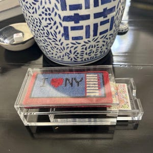 May include: A clear acrylic sliding box containing a deck of playing cards. The top of the box features a needlepoint insert with a blue background, a red heart, and the text I heart NY, bordered by a red frame and a white and blue striped edge.