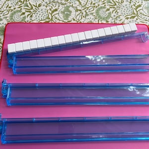 May include: A set of four translucent blue plastic Mahjong racks arranged on a pink surface. One rack holds a row of white tiles, while the others are empty. A small portion of a tile with the word JOKER and the number 8 is visible at the bottom.