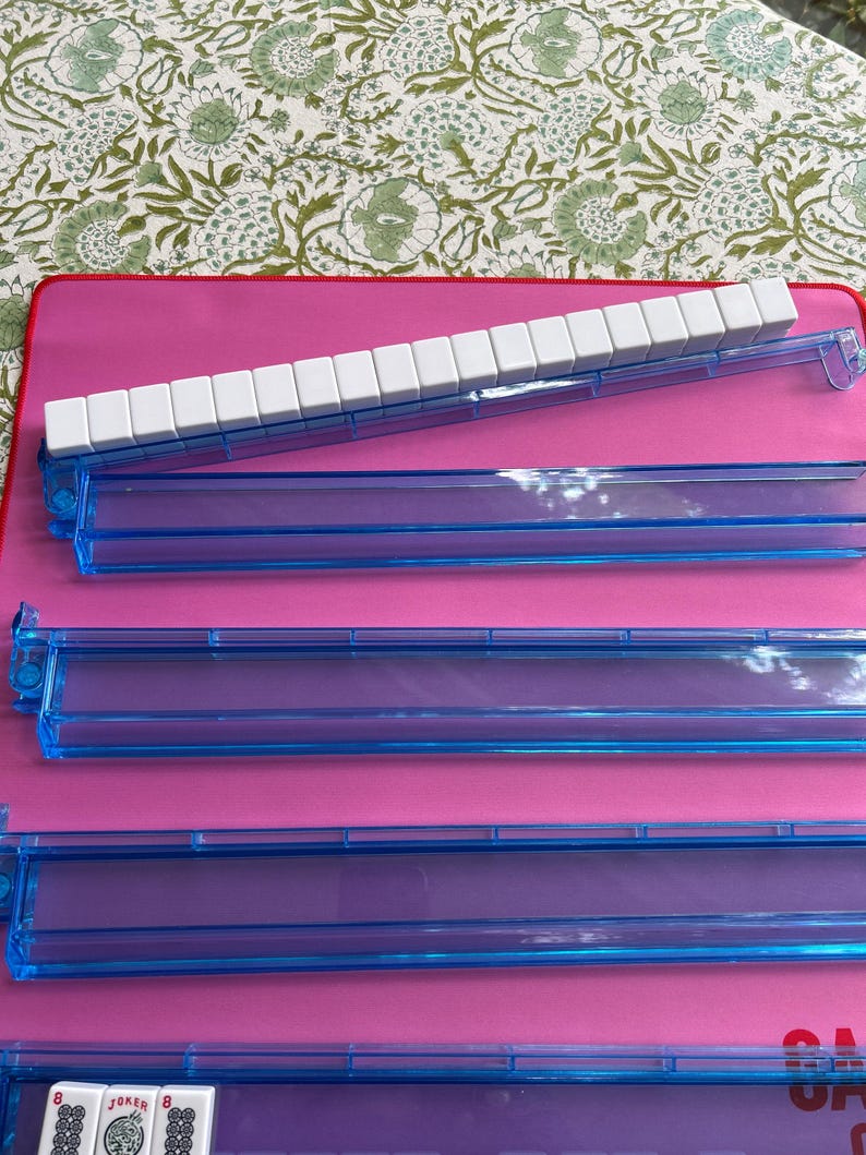 May include: A set of four translucent blue plastic Mahjong racks arranged on a pink surface. One rack holds a row of white tiles, while the others are empty. A small portion of a tile with the word JOKER and the number 8 is visible at the bottom.