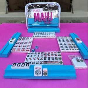 May include: A Mahjong set with white tiles, blue racks, and dice, arranged on a pink surface. The set includes a clear carrying case with the text "MAHJ to go..." in pink and blue.