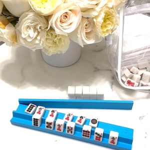 Puede incluir: Un estante azul para fichas de Mahjong sostiene fichas blancas con s&iacute;mbolos rojos y negros. Se ven fichas adicionales, junto con un ramo de rosas blancas y amarillas en un jarr&oacute;n blanco. Una bolsa de pl&aacute;stico transparente contiene m&aacute;s fichas.