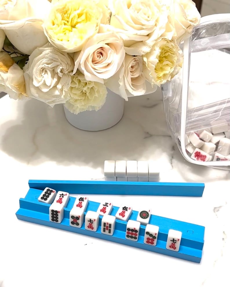 Puede incluir: Un estante azul para fichas de Mahjong sostiene fichas blancas con s&iacute;mbolos rojos y negros. Se ven fichas adicionales, junto con un ramo de rosas blancas y amarillas en un jarr&oacute;n blanco. Una bolsa de pl&aacute;stico transparente contiene m&aacute;s fichas.