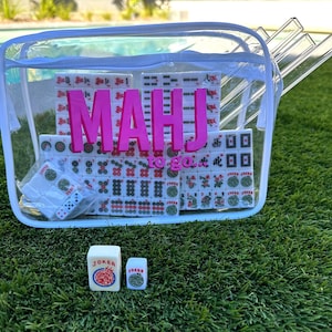 Pode incluir: Uma sacola de plástico transparente contendo um conjunto de Mahjong. O conjunto inclui peças brancas com marcações vermelhas e verdes, dados e racks. As palavras "MAHJ" e "to go" estão em letras rosa nas peças. Duas peças brancas adicionais com a palavra "JOKER" estão na frente da sacola.