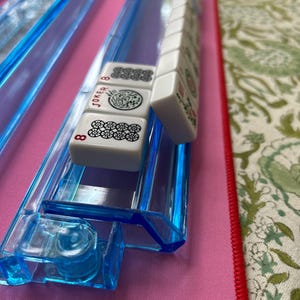 May include: A set of white Mahjong tiles with black and red markings, including a Joker tile and an 8 tile, resting on a translucent blue plastic rack. The tiles feature various traditional symbols and characters printed on their front surfaces.