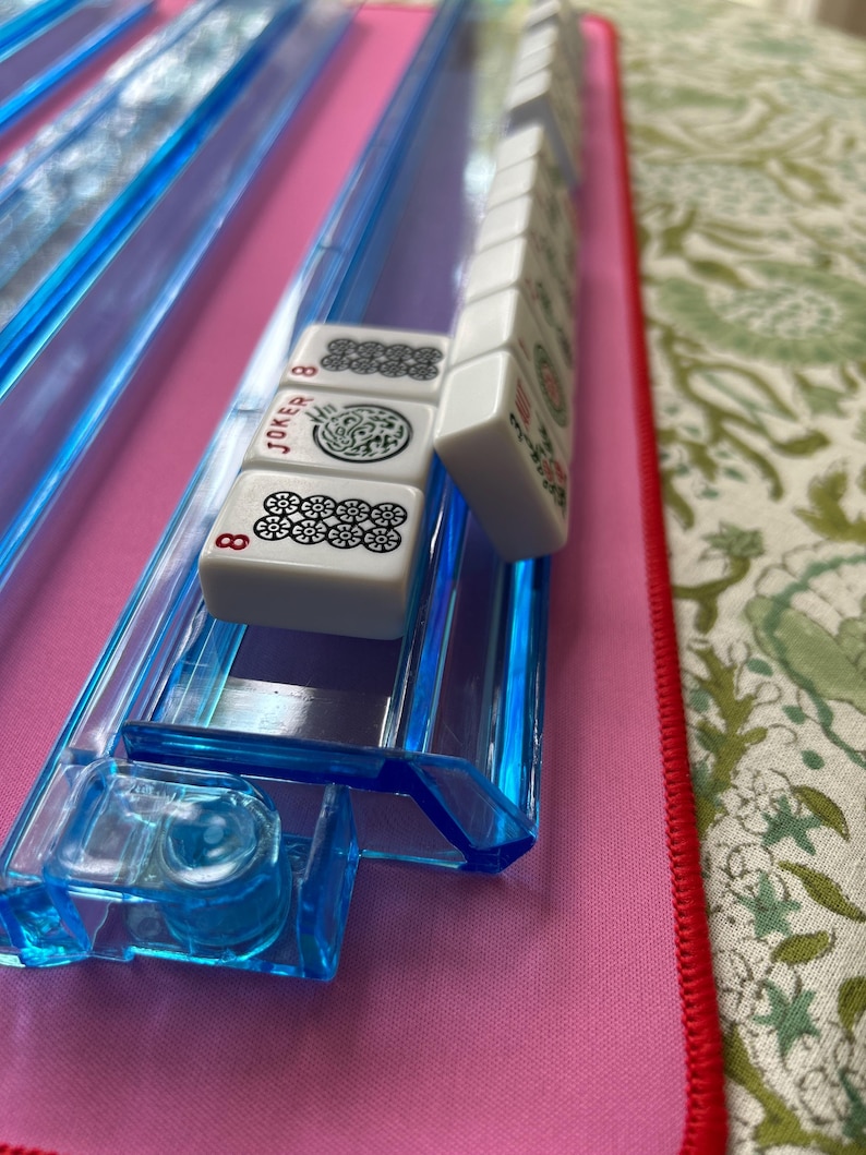 May include: A set of white Mahjong tiles with black and red markings, including a Joker tile and an 8 tile, resting on a translucent blue plastic rack. The tiles feature various traditional symbols and characters printed on their front surfaces.