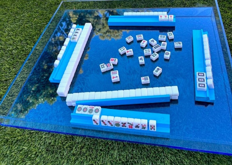 May include: A Mahjong set with blue tile holders and a blue playing surface. The tiles are white with various symbols and characters. The set is arranged on a clear, square surface, with green grass in the background.