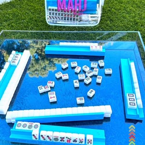 Puede incluir: Un juego de Mahjong con fichas azules y blancas, dispuesto sobre una superficie azul reflectante. El juego incluye estantes azules para fichas y una funda de transporte transparente con la palabra "MAHJ" en letras rosas. El texto "now available!!" est&aacute; en la parte superior.