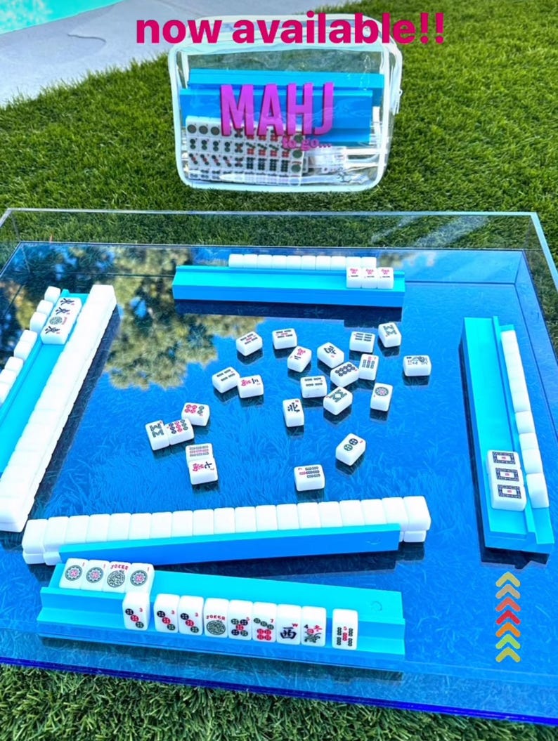 Puede incluir: Un juego de Mahjong con fichas azules y blancas, dispuesto sobre una superficie azul reflectante. El juego incluye estantes azules para fichas y una funda de transporte transparente con la palabra "MAHJ" en letras rosas. El texto "now available!!" est&aacute; en la parte superior.