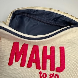 Puede incluir: Una bolsa de lona blanca con cremallera con un dise&ntilde;o bordado en rosa que dice "MAHJ to go..."