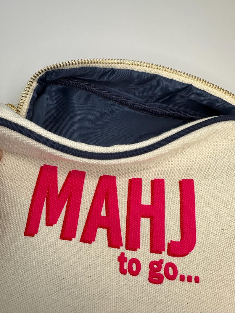 Puede incluir: Una bolsa de lona blanca con cremallera con un dise&ntilde;o bordado en rosa que dice "MAHJ to go..."