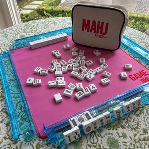 May include: A Mahjong game set featuring a bright pink play mat with blue plastic tile racks, white tiles with black and red symbols, and a beige zippered storage bag with the red text MAHJ to go and GAME ON printed on the mat surface.
