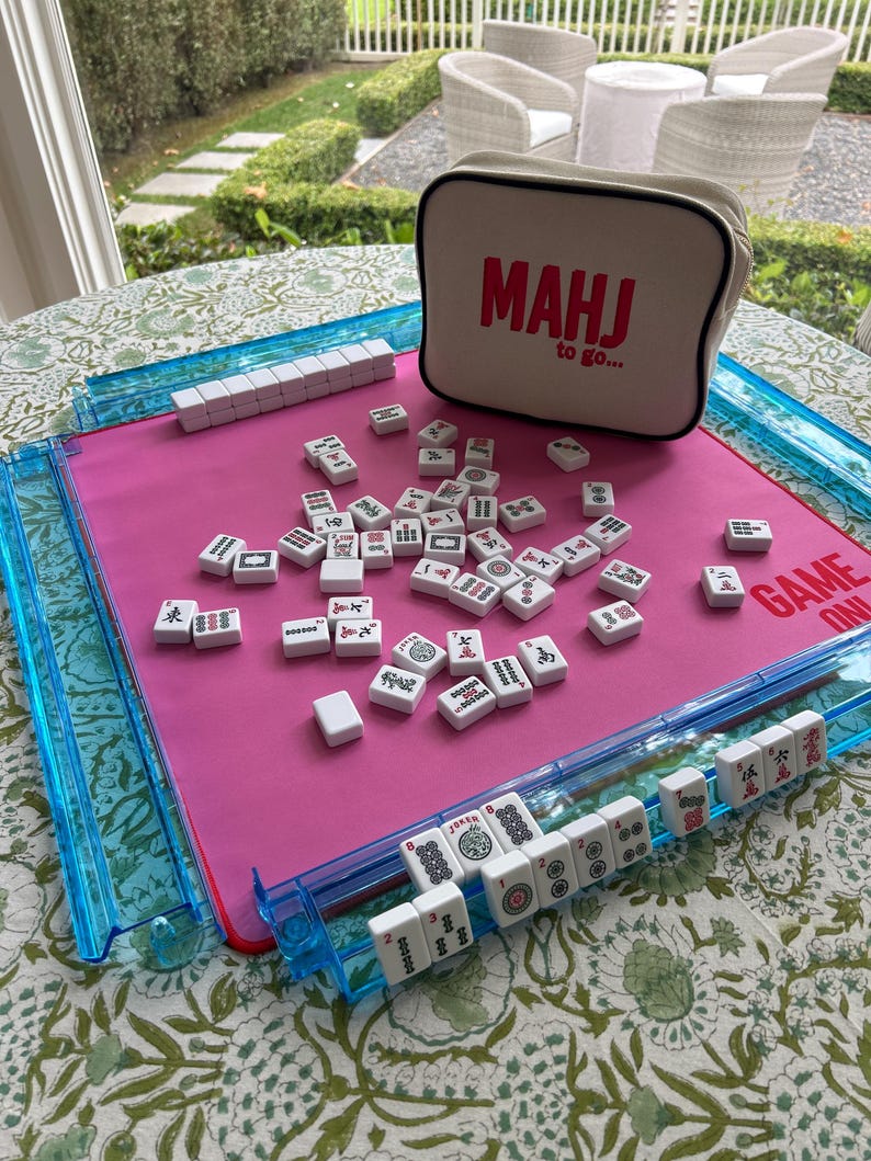 May include: A Mahjong game set featuring a bright pink play mat with blue plastic tile racks, white tiles with black and red symbols, and a beige zippered storage bag with the red text MAHJ to go and GAME ON printed on the mat surface.