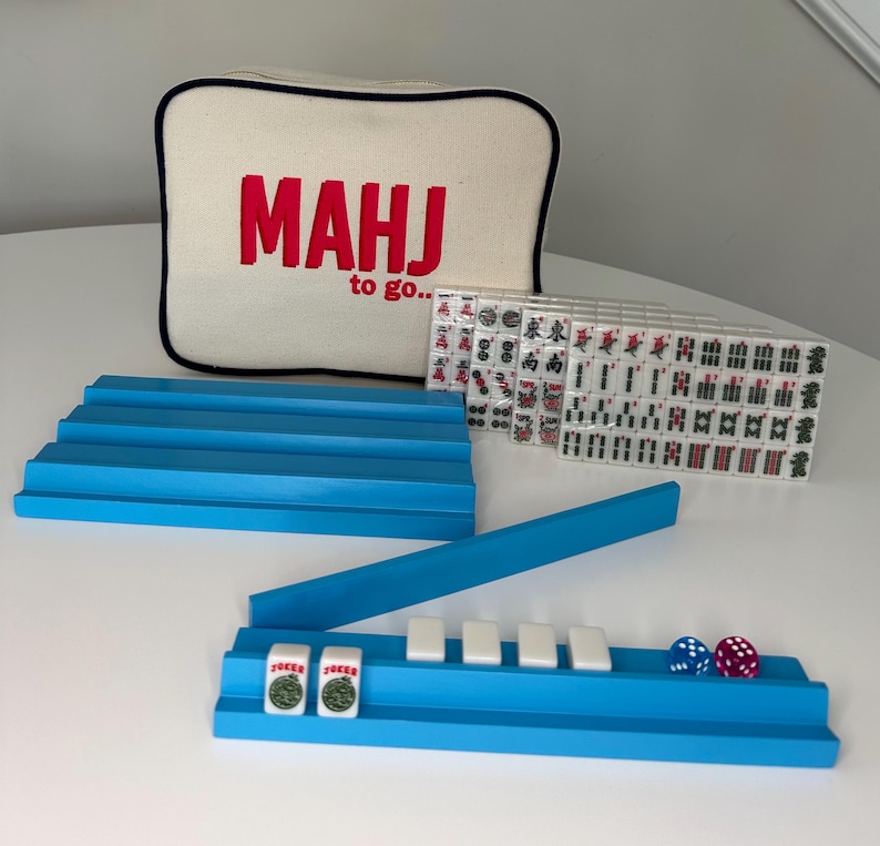 Puede incluir: Un juego de Mahjong con una bolsa de lona blanca que dice "Mahj to go..." en letras rojas. El juego incluye bastidores azules, fichas blancas y dados rojos y azules.