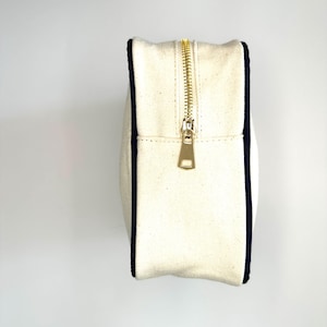 Puede incluir: Una bolsa de lona blanca con cremallera y ribete azul marino. La cremallera es de metal dorado.