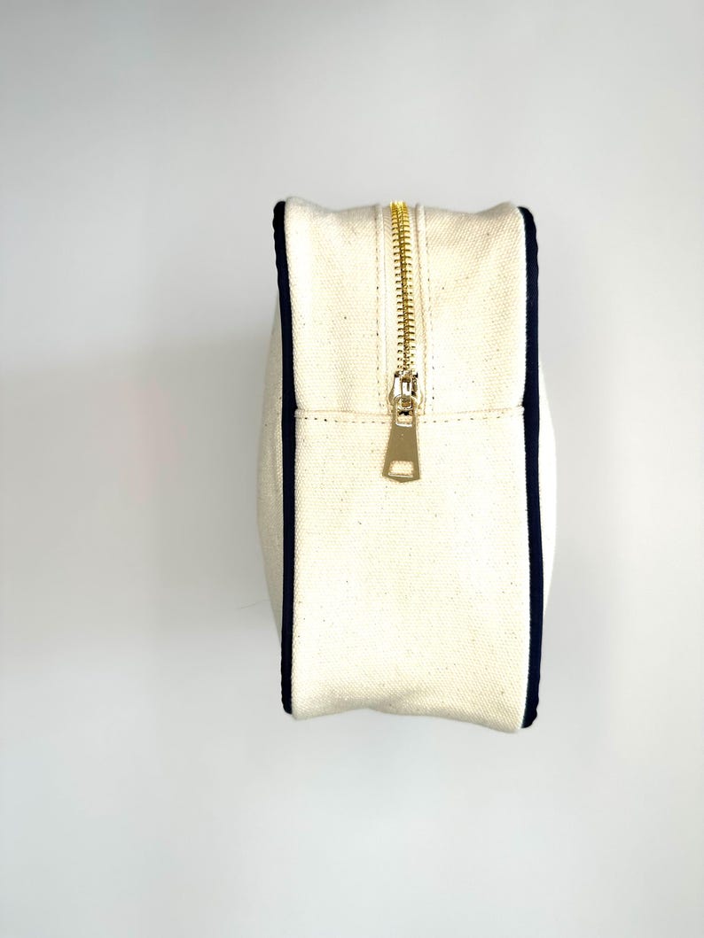 Puede incluir: Una bolsa de lona blanca con cremallera y ribete azul marino. La cremallera es de metal dorado.